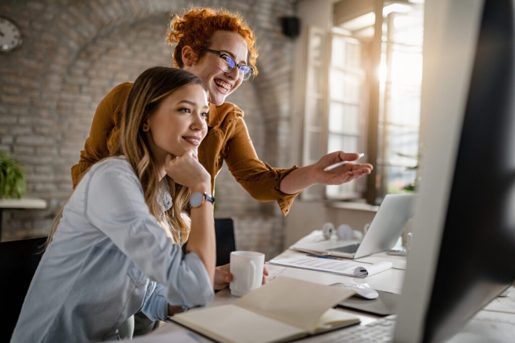 Sobre Empresas 4 empresarias felizes lendo um e mail no computador enquanto trabalham juntas no escritorio o foco esta na mulher ruiva scaled e1702193271993